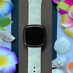 May include: A light green smartwatch band with a sea turtle pattern. The band is surrounded by colorful flowers, seashells, and small green turtle illustrations. The watch face is black.