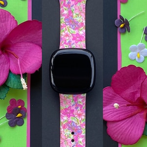 K&ouml;nnte beinhalten: Ein pinkfarbenes und wei&szlig;es Armband mit floralem Muster und einem schwarzen Zifferblatt. Das Armband besteht aus Silikon und hat einen Blumendruck. Das Zifferblatt ist rund und hat eine schwarze L&uuml;nette.