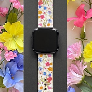 May include: A smartwatch with a white floral patterned band featuring red, blue, yellow, and orange flowers. The watch is surrounded by colorful flowers, including pink, yellow, and blue blooms, creating a vibrant display.