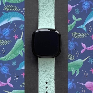 May include: A light green silicone watch band with a white narwhal and seahorse design. The band is compatible with Apple Watch.
