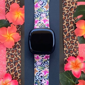 Puede incluir: Un reloj inteligente negro con una banda de silicona con estampado floral y de leopardo. La banda tiene flores rosas, blancas y verdes sobre un fondo blanco con un estampado de leopardo.