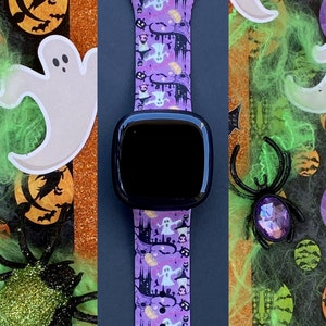 May include: A purple and black watch band with a Halloween theme. The band features ghosts, pumpkins, and haunted houses. The watch face is black and the band is made of silicone.