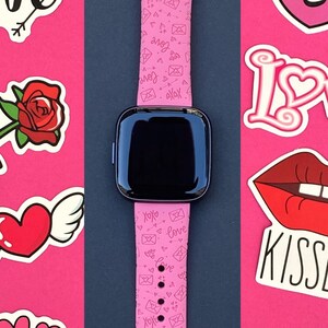 May include: Pink smartwatch with a pink band featuring a repeating pattern of hearts and envelopes. The band has a black buckle. The watch face is black and has a white digital clock.