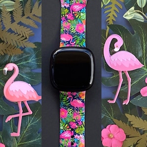 May include: A black smartwatch with a pink and green tropical print watch band featuring pink flamingos and palm leaves.