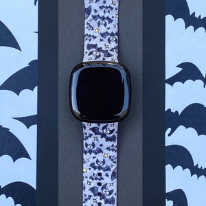 Puede incluir: Un reloj inteligente negro con una banda de color morado y negro con estampado de murci&eacute;lagos. La banda tiene peque&ntilde;as estrellas doradas.