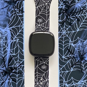 Op de afbeelding: Een zwarte smartwatch met een witte spinnenweb-patroonband. De band heeft kleine spin-silhouetten erop. Het horloge bevindt zich op een witte achtergrond met een zwart en blauw spinnenweb-patroon aan beide kanten.