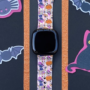 以下が含まれることがあります： ピンクとオレンジのハロウィーンをテーマにしたバンドが付いた黒いスマートウォッチ。バンドには、猫、カボチャ、コウモリ、キャンディーコーンが繰り返しパターンで描かれています。