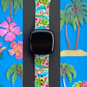 May include: A black smartwatch with a tropical floral print band. The band features pink hibiscus flowers, palm trees, and blue water. The watch face is black and the band is made of silicone.