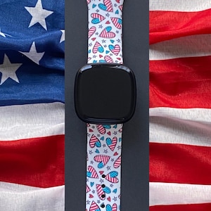 May include: A black smartwatch with a white strap patterned with red, white, and blue hearts and stars. The background features the American flag.