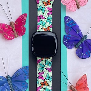 May include: A black smartwatch with a green, white, and pink butterfly patterned band. The band has a white background with small white dots and colorful butterflies.