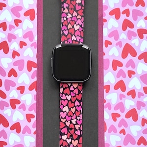 K&ouml;nnte beinhalten: Eine Smartwatch mit schwarzem Bildschirm und einem rosa Armband mit Herzmuster. Das Armband zeigt ein sich wiederholendes Muster aus roten, rosa und wei&szlig;en Herzen auf schwarzem Hintergrund. Die Uhr wird auf einer schwarzen Oberfl&auml;che mit einem rosa Herzmuster-Hintergrund pr&auml;sentiert.