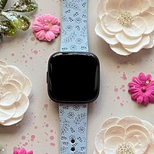 K&ouml;nnte beinhalten: Ein hellblaues Armband mit einem wei&szlig;en Blumenmuster. Das Armband ist an einer Smartwatch mit schwarzem Zifferblatt.