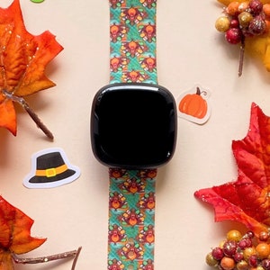 K&ouml;nnte beinhalten: Ein t&uuml;rkisfarbenes und orangefarbenes Armband mit einem schwarzen Zifferblatt. Das Armband zeigt ein sich wiederholendes Muster aus Truth&auml;hnen mit Pilgerh&uuml;ten. Die Uhr ist von Herbstdekorationen umgeben, darunter Bl&auml;tter, K&uuml;rbisse und Beeren.