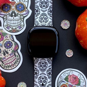 May include: A white and black watch band with a sugar skull pattern. The band is on a black surface with several small, sparkly, silver decorations. There are also several orange pumpkins and fall leaves on the surface.