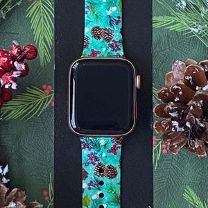 May include: A teal and green Apple Watch band with a pine cone and berry pattern. The band is on a black background with pine branches and red berries.