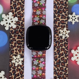 May include: A black smartwatch with a colourful Christmas lights and snowflakes patterned band. The band is on a brown leopard print background.