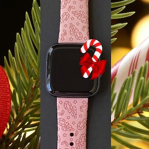 May include: Pink Apple Watch band with a candy cane and star pattern. The band is shown on a black background with a candy cane and a red bow.