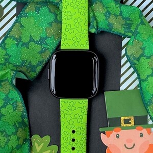 May include: A green silicone watch band with a shamrock pattern. The band is on a black surface with a green and gold ribbon with a shamrock pattern.