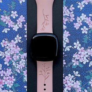 May include: A pink silicone Apple Watch band with a floral design. The band has a silver buckle and is shown on a blue background with a floral pattern.