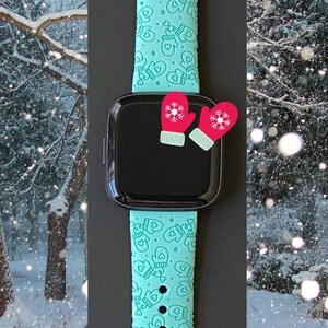 May include: A teal smartwatch band with a pattern of mittens and hearts. The watch face is black, and two red mitten-shaped charms are attached. The background shows a snowy winter scene.