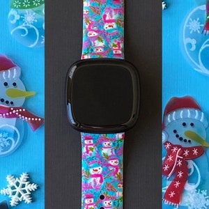 K&ouml;nnte beinhalten: Ein pinkfarbenes, blaues und wei&szlig;es Armband mit einem schwarzen Zifferblatt. Das Armband hat ein sich wiederholendes Muster aus Schneem&auml;nnern.