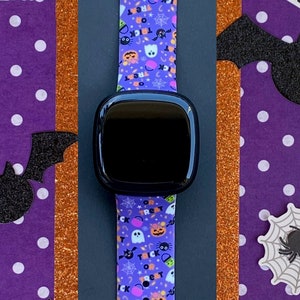 Puede incluir: Una banda de Apple Watch con estampado morado y negro con una esfera de reloj negra. La banda presenta una variedad de im&aacute;genes con tem&aacute;tica de Halloween, incluidos fantasmas, calabazas, murci&eacute;lagos y ma&iacute;z dulce. El reloj est&aacute; rodeado de decoraciones de Halloween, que incluyen un gato negro, una telara&ntilde;a y una calabaza.