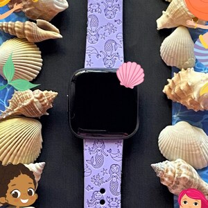 May include: A purple silicone Apple Watch band with a mermaid pattern. The band features a variety of mermaids, seashells, and starfish. The band is surrounded by seashells and a blue and white background.