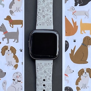 May include: A smartwatch with a black screen and a light gray band. The band is decorated with a pattern of smiling faces, flowers, and bones. The background features a repeating pattern of various dog breeds in different colors.