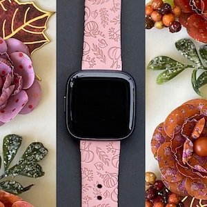 K&ouml;nnte beinhalten: Ein rosafarbenes Apple Watch Armband mit einem Herbst-Design mit K&uuml;rbissen und Bl&auml;ttern. Das Armband wird vor einem Hintergrund aus herbstlichen Dekorationen gezeigt.