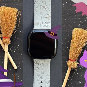 May include: A white watch band with a Halloween theme. The band has a white background with black text and illustrations of witches, brooms, and cats. The text on the band says "Basic Witch".