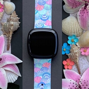 K&ouml;nnte beinhalten: Ein hellblaues, rosa und wei&szlig;es Armband mit Muschel-Muster. Das Armband ist aus Silikon und f&uuml;hlt sich angenehm weich an. Es ist kompatibel mit der Apple Watch Series 4 und neuer.