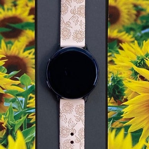May include: A black smartwatch with a light pink band featuring a sunflower design. The band is made of a soft material and has a series of holes for adjustment. The watch face is black and round.