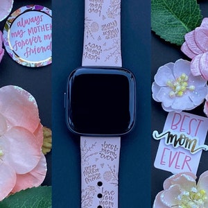 May include: A pink Apple Watch band with a floral design and gold lettering. The band has the words "best mom ever", "love you mom", "always my mother forever my friend", and "cherished always you're the best mom ever" printed on it.