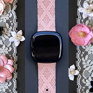 Puede incluir: Una banda de reloj inteligente rosa con un delicado patr&oacute;n floral. La banda tiene una hebilla plateada y se muestra sobre un fondo negro con ribete de encaje.