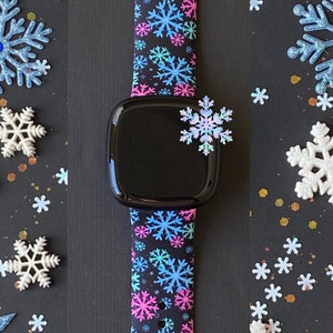 K&ouml;nnte beinhalten: Ein schwarzes Smartwatch-Armband mit einem Schneeflockenmuster in Pink, Blau und Wei&szlig;. Das Armband ist aus Silikon und kompatibel mit Apple Watch.