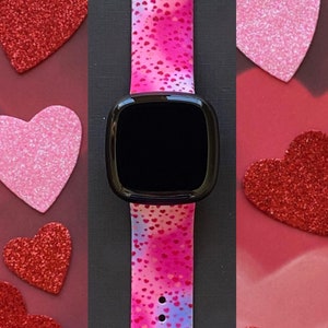 May include: A pink and blue smartwatch band with a white background and small pink hearts printed on it. The band is made of silicone and has a black watch face.