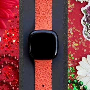 May include: A bright orange Apple Watch band with a repeating pattern of sugar skulls. The band is shown on a watch with a black face. The background is red with white flowers and green beads.