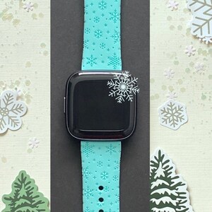 May include: A turquoise blue Apple Watch band with a snowflake pattern. The band is made of silicone and has a smooth finish. The watch band is shown on a white background with snowflakes and Christmas trees.