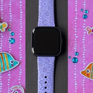 May include: A purple watch strap with a white and blue fish pattern. The strap is on a pink background with colourful fish stickers.
