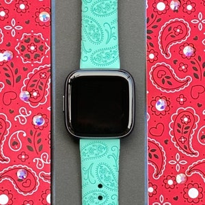 May include: A turquoise green Apple Watch band with a paisley pattern. The band is in a black box with a red and white paisley patterned background.