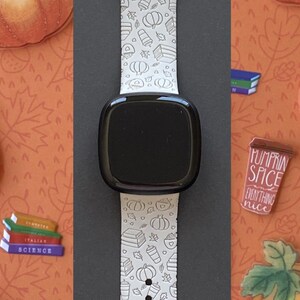 May include: A white smartwatch band with a black watch face, featuring a repeating pattern of pumpkins, books, and coffee cups. The band is surrounded by pumpkin and leaf illustrations on an orange background. The words "Pumpkin Spice Everything Nice" are on a small brown cup.