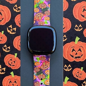 May include: A black smartwatch with a bright pink, orange, and green patterned band featuring a repeating pattern of jack-o'-lanterns. The band is made of silicone and has a buckle closure.