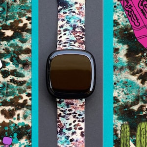 Może przedstawiać: Smartwatch z czarną tarczą i kolorowym paskiem z wzorem czaszki krowy, podkowy, kaktusa i kowbojskich but&oacute;w. Pasek jest wykonany z silikonu i ma zapięcie.