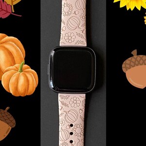 May include: A pink watch band with a fall-themed design. The band features pumpkins, sunflowers, acorns, and leaves.