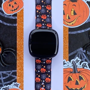 May include: A black smartwatch with a Halloween-themed band. The band features a repeating pattern of orange pumpkins with black faces and white stems, candy corn, and spiderwebs on a black background.