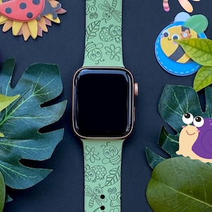 May include: A green Apple Watch band with a white background and a pattern of ladybugs, bees, butterflies, and flowers. The band is on a black background with various cutouts of the same pattern.