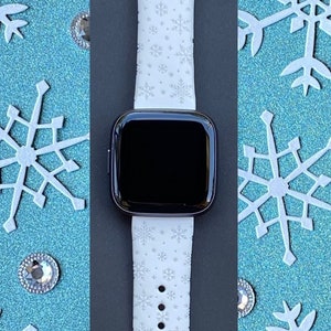 K&ouml;nnte beinhalten: Wei&szlig;es Smartwatch-Armband mit Schneeflockenmuster. Das Armband ist aus Silikon und hat ein schwarzes Zifferblatt.