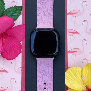May include: A light purple smartwatch band with a floral pattern, set against a pink flamingo-themed background. The band is paired with a black smartwatch face. Decorative pink and yellow flowers with green leaves are placed around the band.