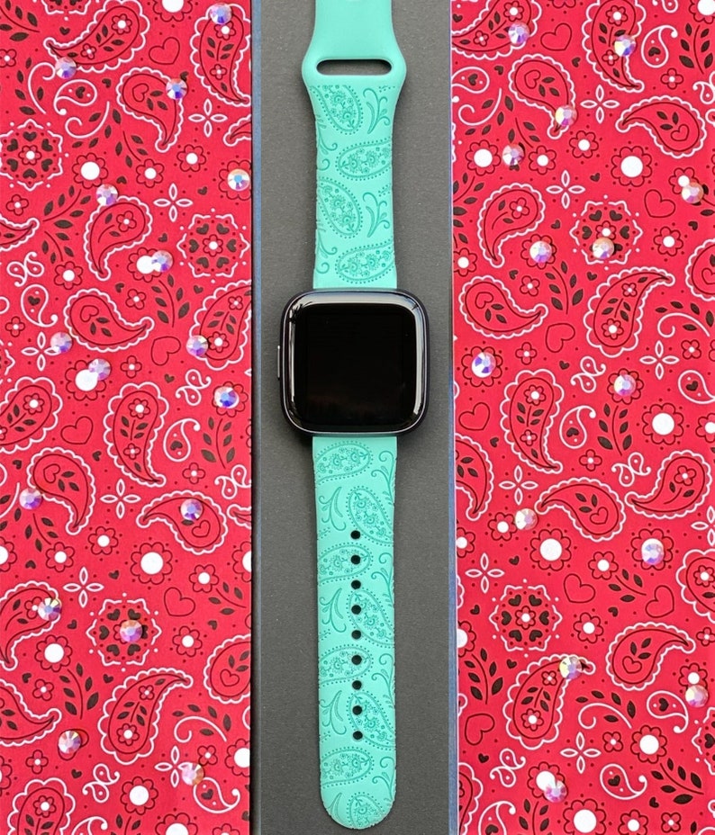 May include: A turquoise green Apple Watch band with a paisley pattern. The band is in a black box with a red and white paisley patterned background.