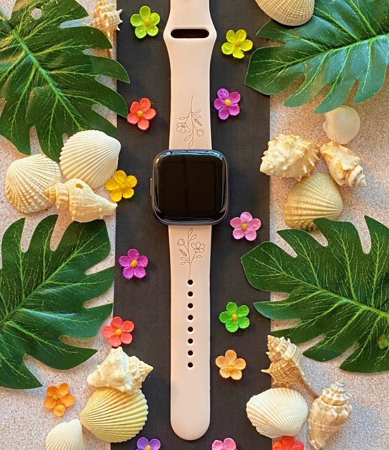 May include: A pink silicone watch band with a floral design. The band has a black watch face and is surrounded by seashells and flowers.